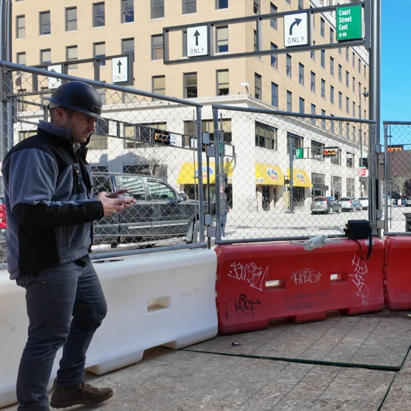 A man in a hard hat walking past a construction fence, phone in hand